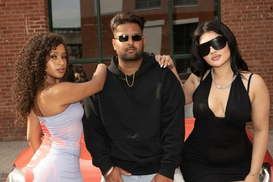 A man in a black hoodie and sunglasses poses between two women in front of a brick wall, behind the scenes at a music video shoot